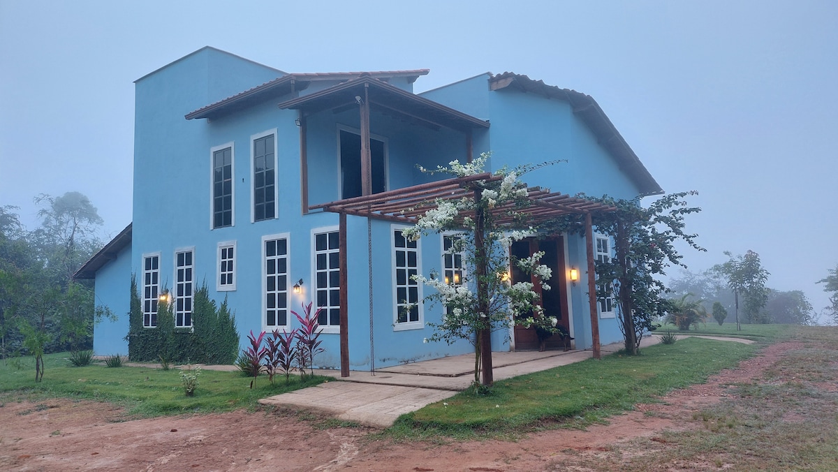 A modern blue house is featured, set against a foggy backdrop. The entrance is framed by a wooden pergola adorned with plants and outdoor lighting. Large windows invite natural light, while a manicured lawn surrounds the property, enhancing the natural scenery.