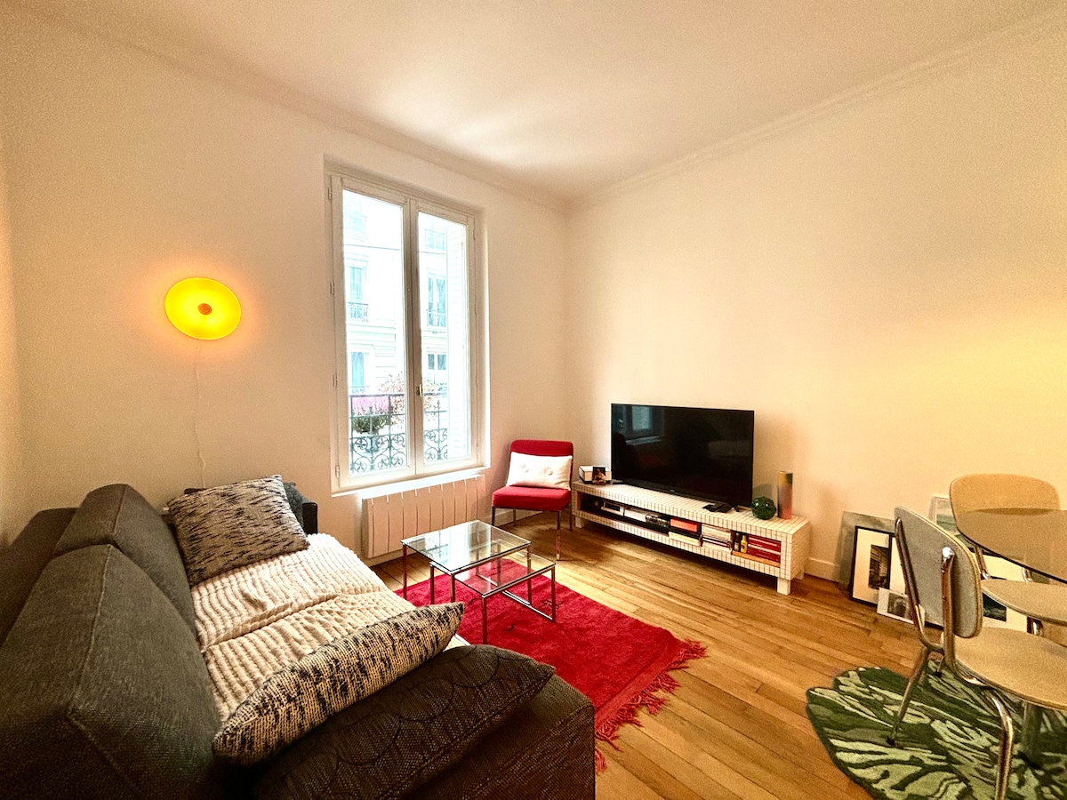 The living area features a cozy gray sofa with a textured throw, a glass coffee table, and a simple red area rug. A modern TV stand with a flat-screen television is positioned against the wall, complemented by a dining table with transparent chairs. Natural light is illuminated through large windows.