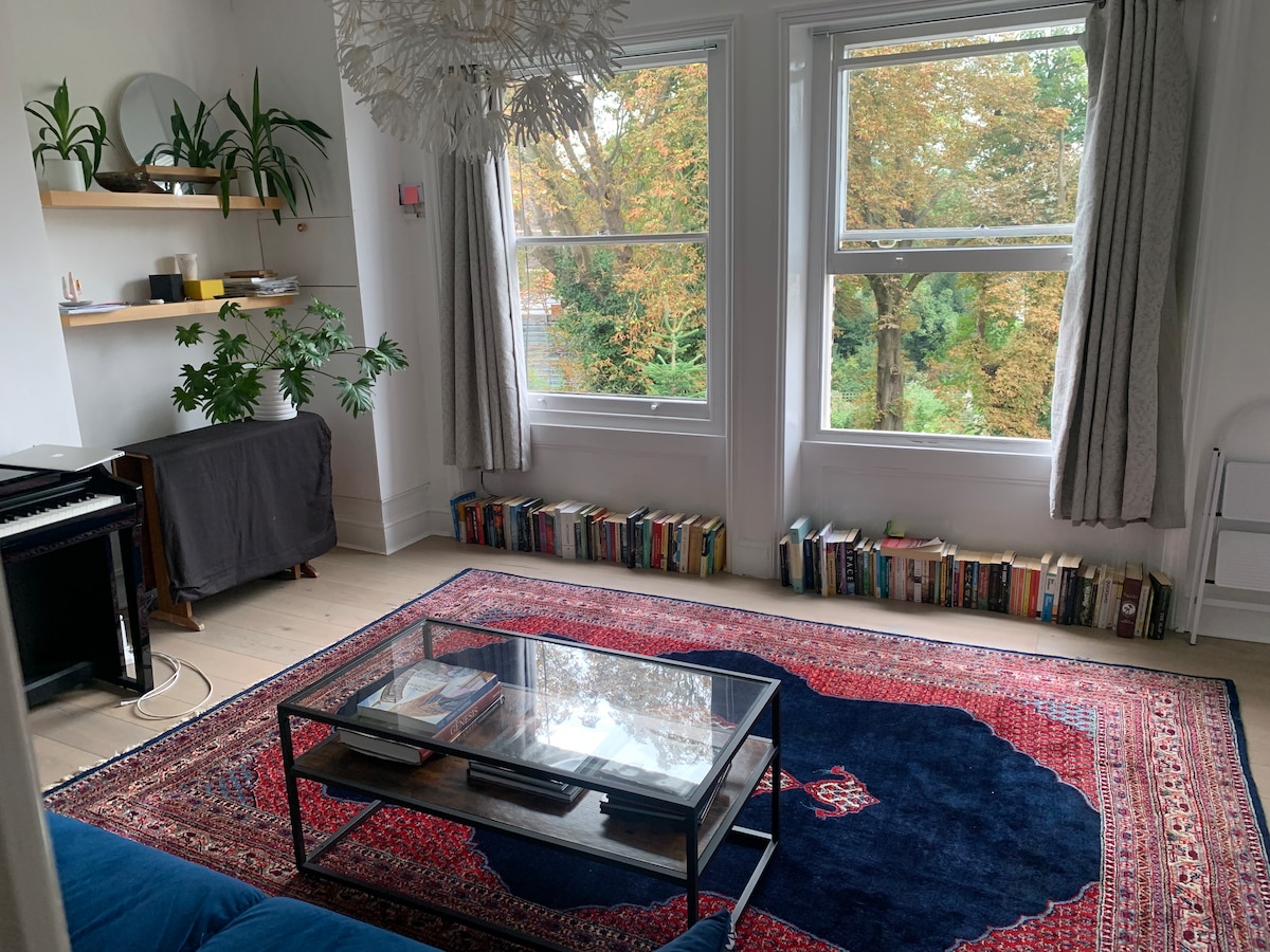 A comfortable living space features large windows framed by soft curtains, allowing natural light to filter in. A glass coffee table sits atop a colorful rug, surrounded by a seating area. Beneath the windows, a collection of books is neatly arranged on a low shelf.