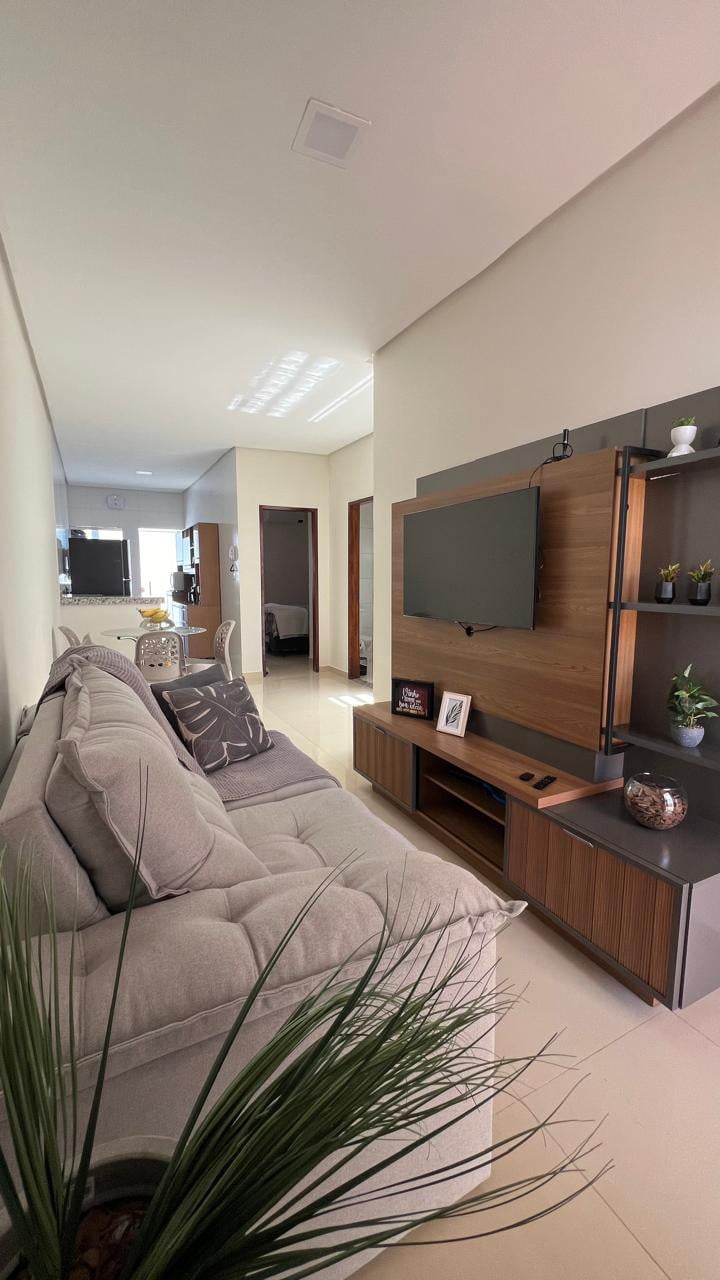 A well-lit living area is seen, featuring a comfortable sofa and a modern entertainment center with a mounted TV. A potted plant is positioned in the foreground, adding a touch of greenery. Light streams in through windows, illuminating the neutral-toned space.