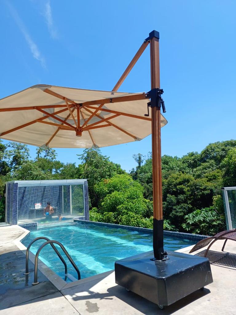 A modern swimming pool is surrounded by lush greenery, with a large umbrella providing shade. The pool features a textured surface and steps leading into the water. A person can be seen enjoying the pool in the background, enhancing the tranquil setting.