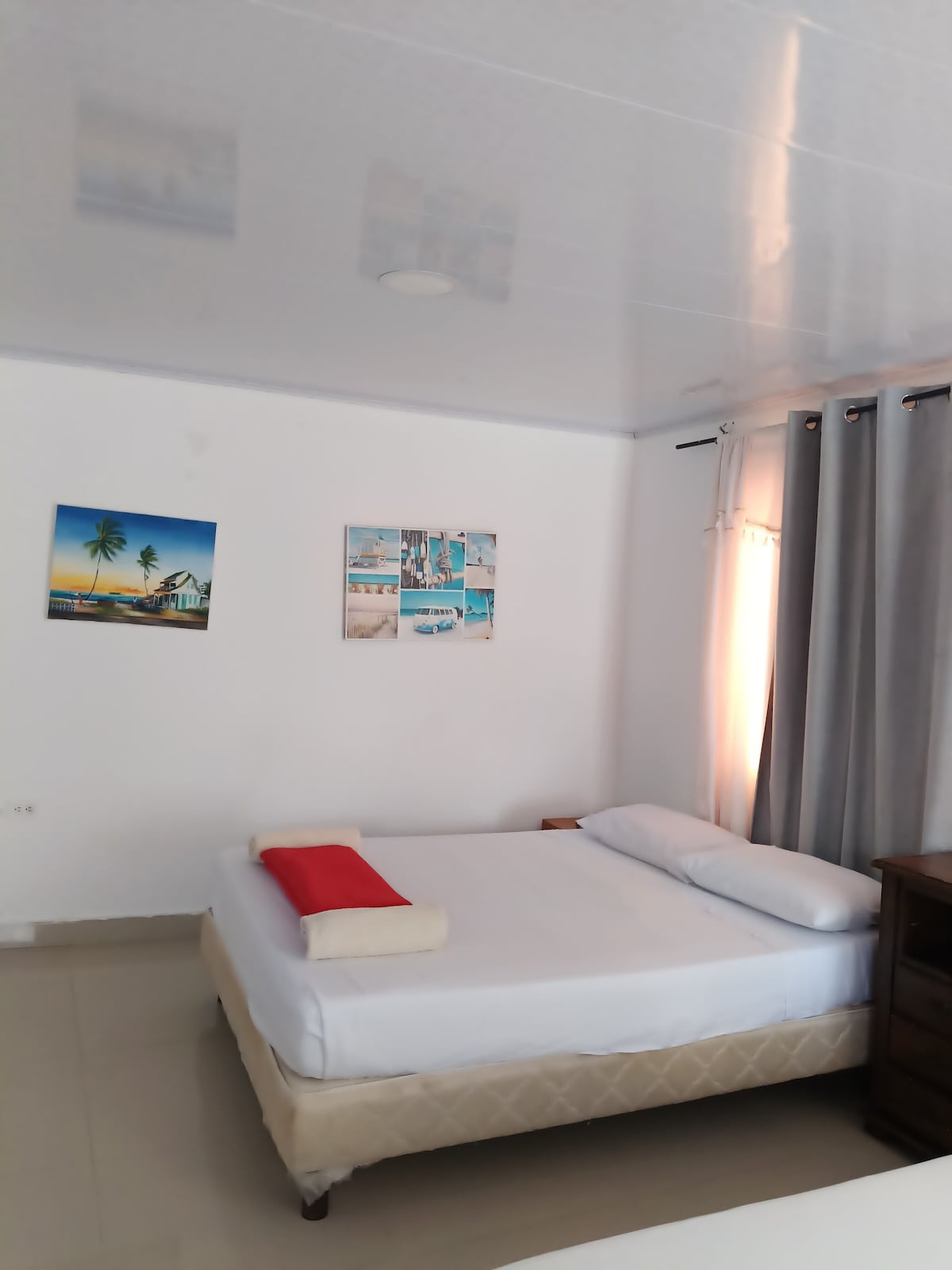 A comfortable sleeping area is featured with a spacious bed dressed in crisp white linens. A decorative red pillow adds a pop of color. Two framed images adorn the walls, complemented by soft gray curtains that filter natural light from a nearby window.
