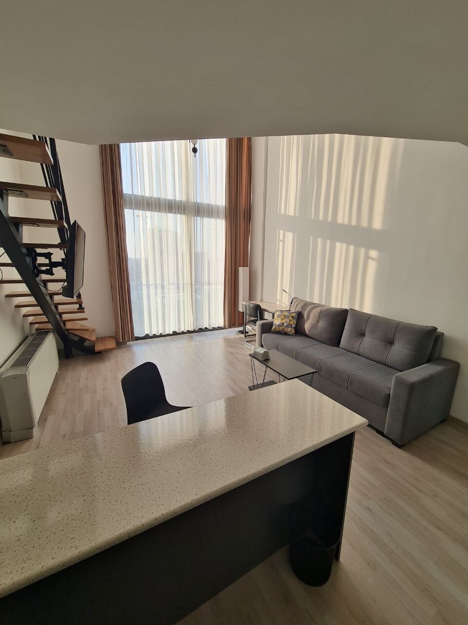 The spacious living area features large windows allowing natural light to fill the room, complemented by soft fabric furnishings. A staircase leads to the upper level. A sleek countertop serves as a barrier between the living space and kitchen area, enhancing the open layout.
