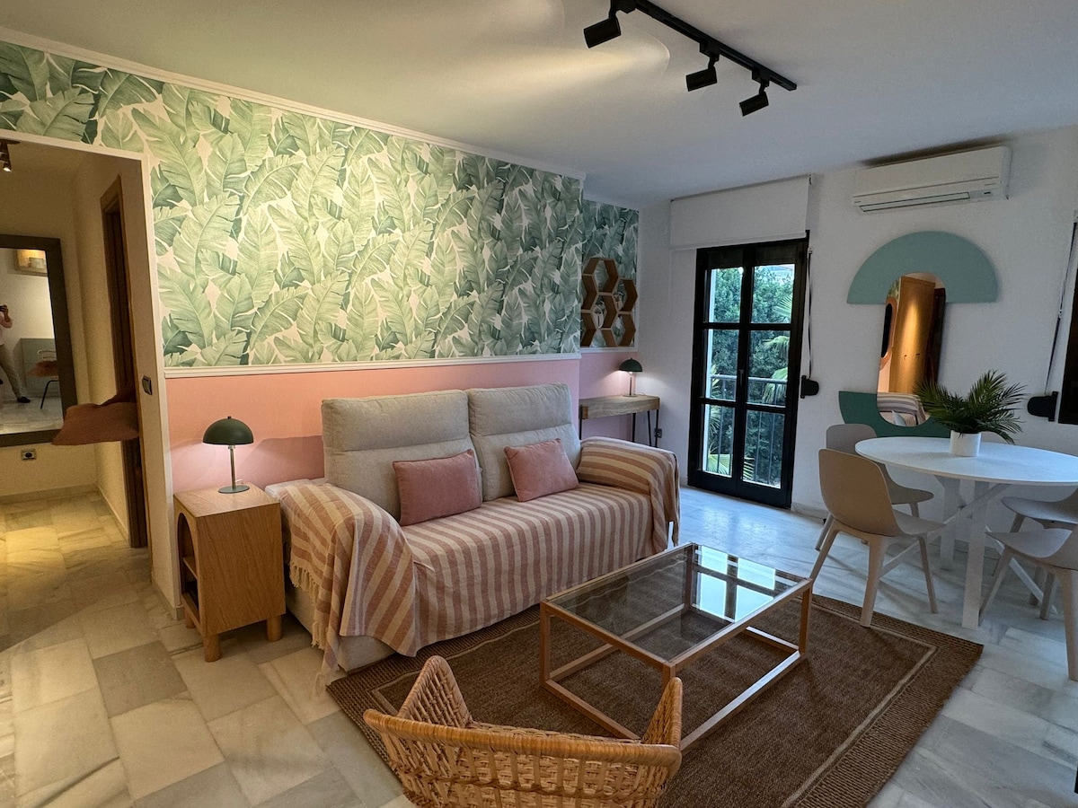 A comfortable living area features a soft sofa with decorative pillows, accented by wallpaper with a lush plant motif. A coffee table sits on a textured rug, accompanied by a light wood dining table and chairs. Large windows allow natural light to fill the space.