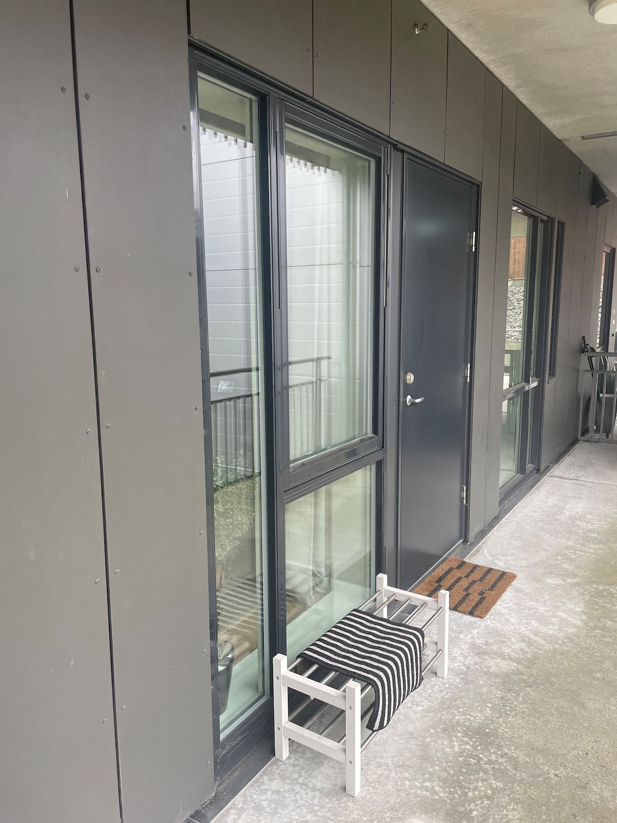 The entrance features large glass doors framed by sleek black panels, allowing natural light to illuminate the area. A small white folding stool is positioned in front of a striped welcome mat, enhancing the entryway's accessibility.