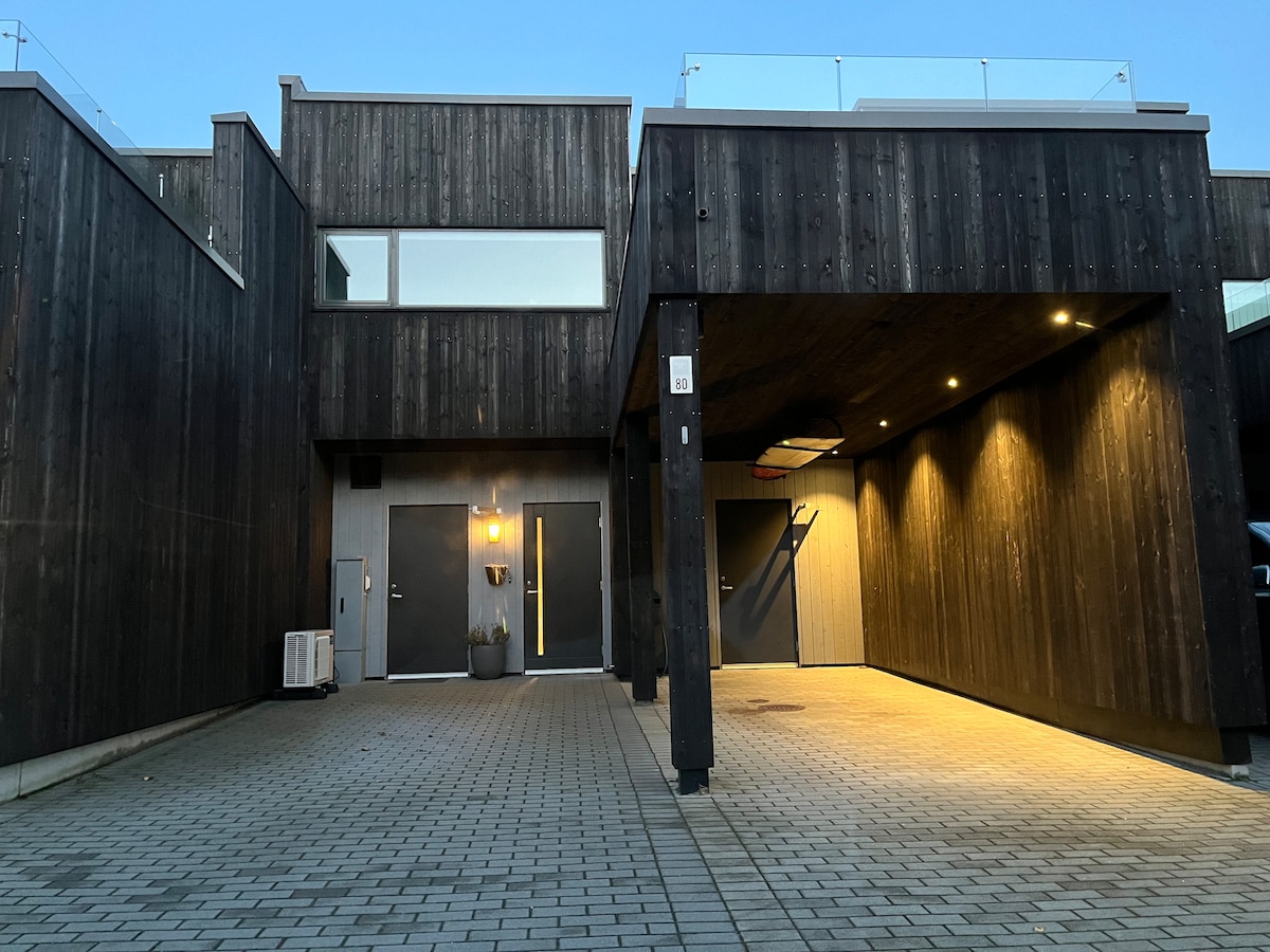 The exterior of the house features a contemporary wooden facade with large windows and a covered entrance. The paved driveway leads to dual entry doors, illuminated by soft exterior lighting. A minimalistic planter adds a touch of greenery beside the entrance.