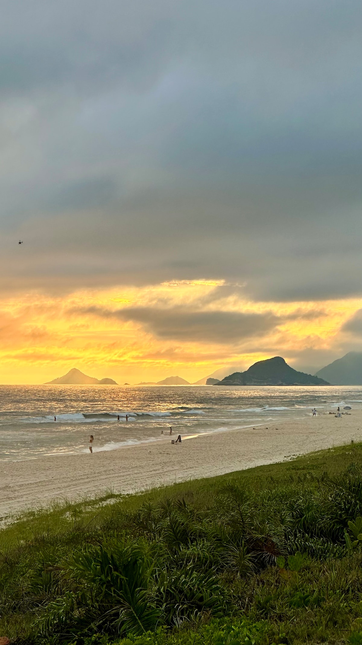 A serene beach scene captures the sunset over the ocean, with soft waves lapping against the shore. Silhouettes of surfers are seen in the water, while distant mountains rise against the cloudy sky. Lush green vegetation is present in the foreground, adding to the natural beauty.