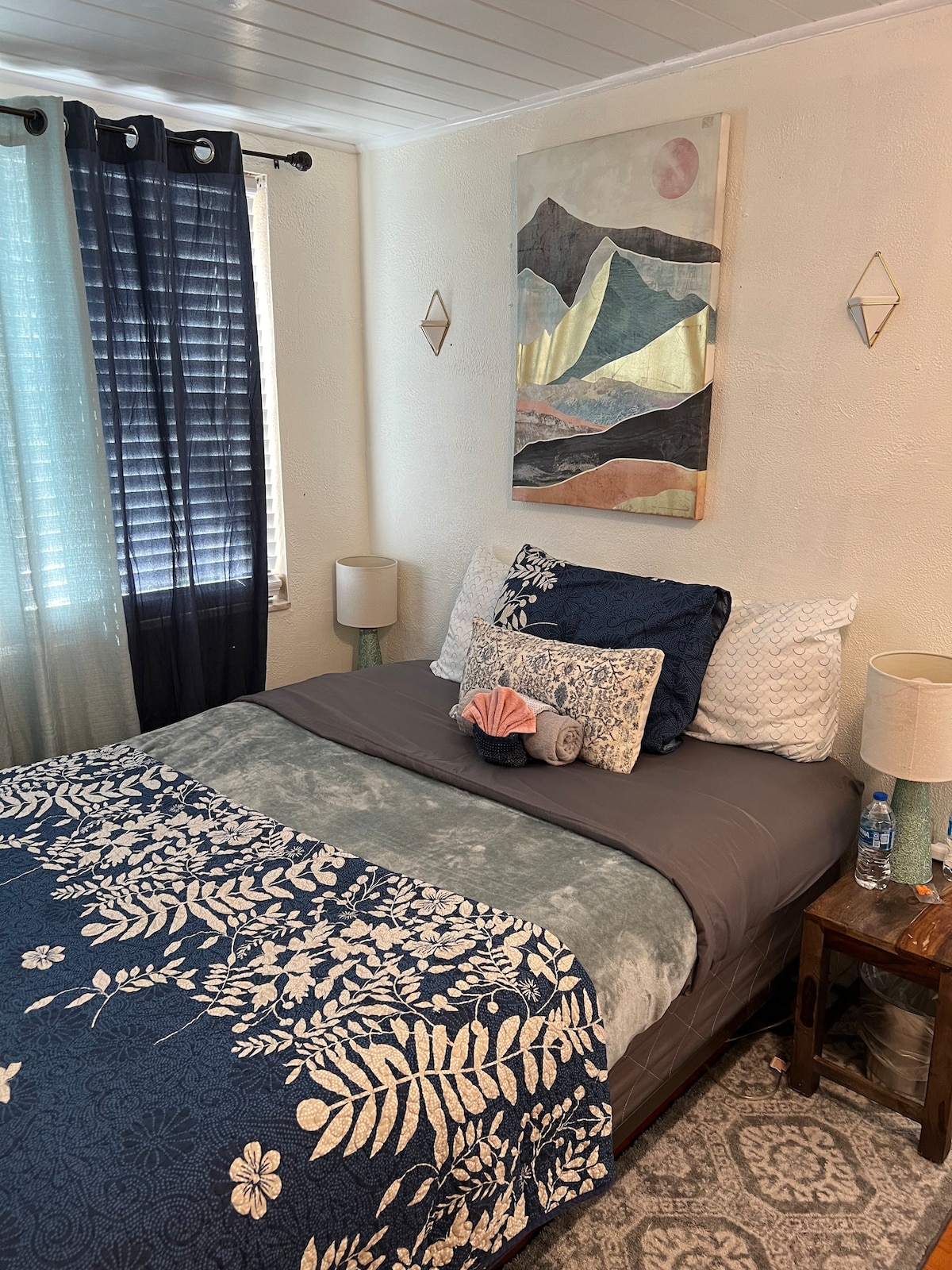 A cozy room is showcased with a full-size bed adorned in blue and gray linens. Decorative pillows, including a seashell, add a personal touch. Light filters through sheer curtains, and two bedside lamps offer illumination. Artwork with earthy tones hangs on the wall, enhancing the serene atmosphere.