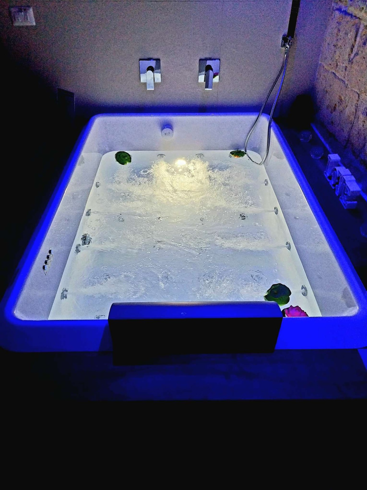 A spacious jacuzzi is featured, highlighted by soft lighting and water jets. Floral accents float on the surface, and the surrounding area is designed for relaxation, with modern fixtures visible on the wall nearby.
