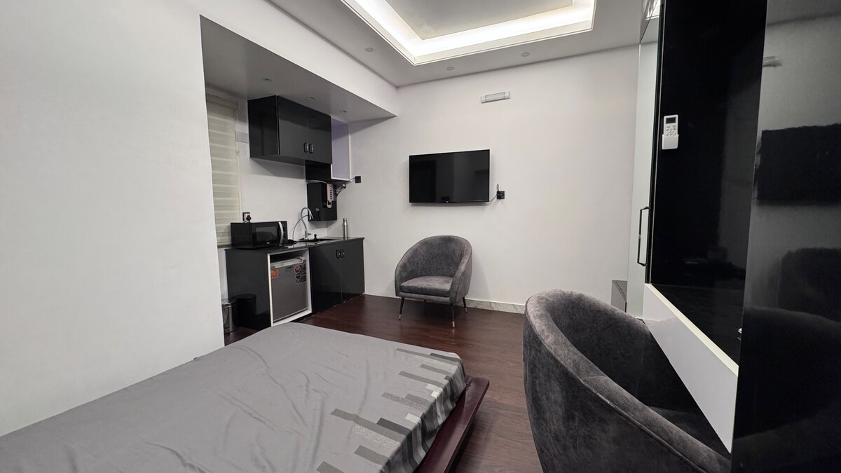 A modern studio is displayed, featuring a spacious bed with a neutral-colored sheet. To one side, a kitchenette with sleek cabinetry and appliances is visible. Two plush, curved chairs complement the contemporary design, and a flat-screen TV is mounted on the wall, enhancing the minimalist aesthetic.