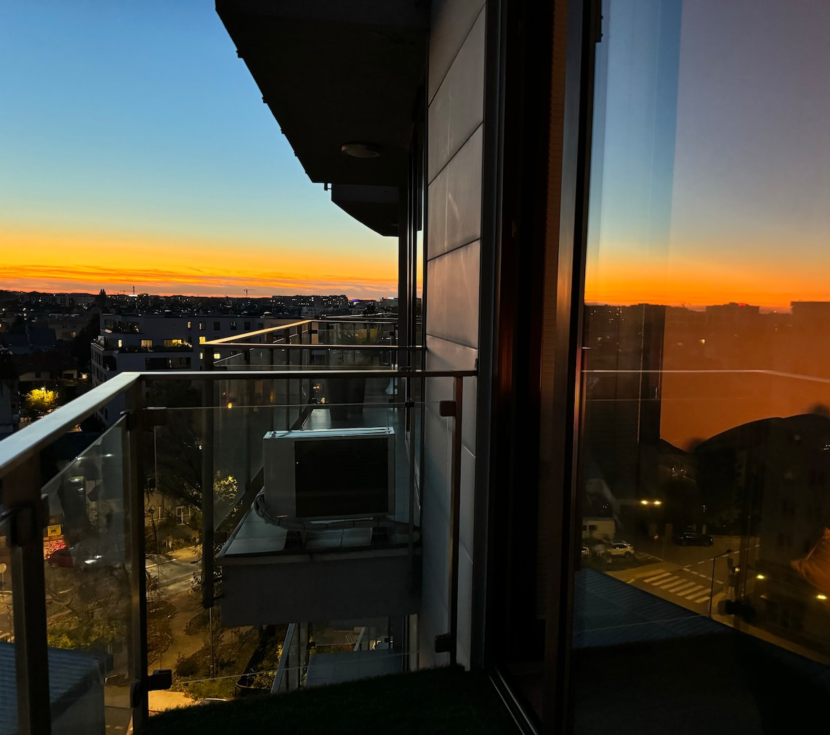 A balcony view captures a serene sunset horizon, blending warm orange and blue tones. Reflections of the surrounding area are visible on the glass, enhancing the sense of spaciousness. The image illustrates the peaceful atmosphere of the evening, framed by sleek balcony railings.