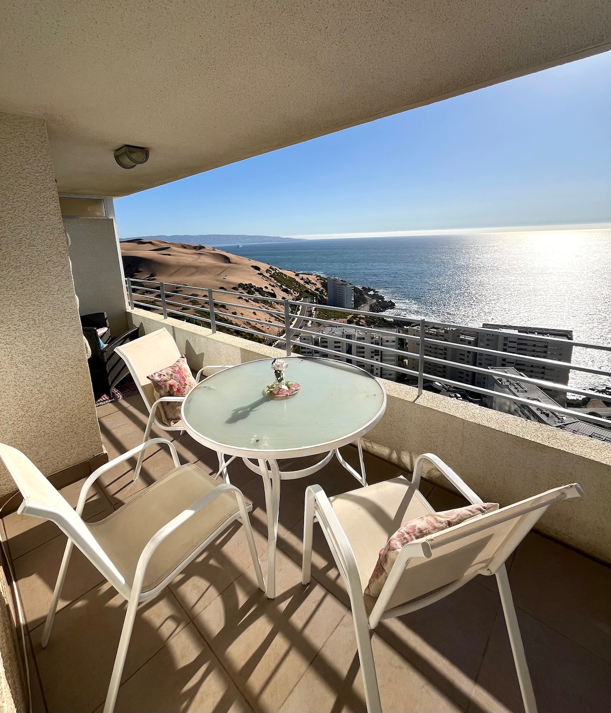 A balcony space features a round glass table surrounded by four white chairs. The view includes the expansive ocean and a distant coastline. Natural light illuminates the area, emphasizing the serene atmosphere. The railing provides safety while maintaining an unobstructed view.