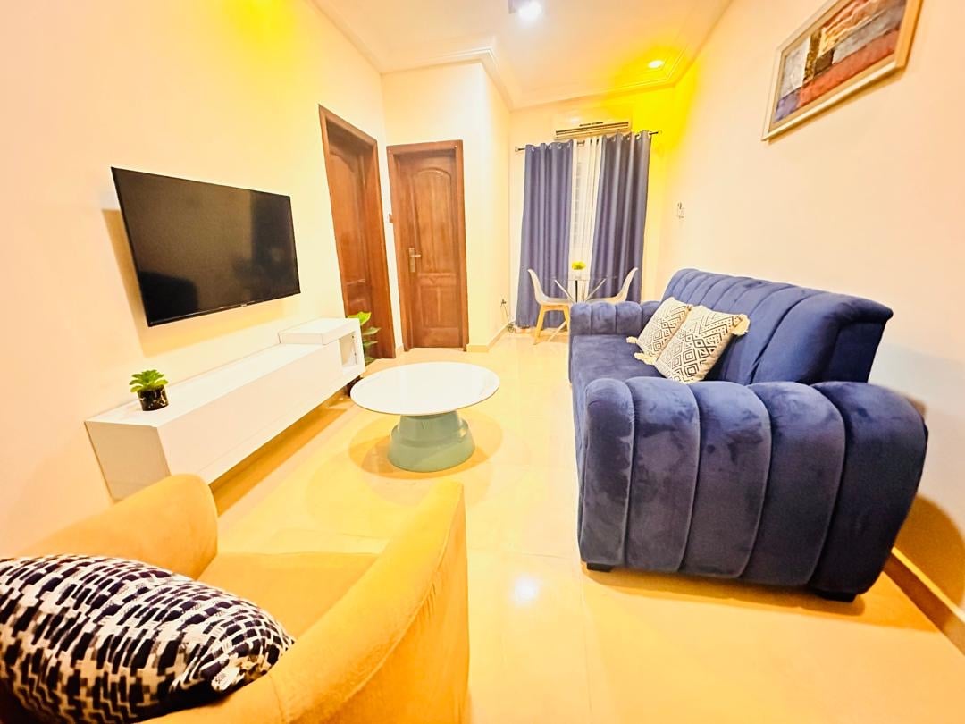 A comfortable living area features a blue sofa and an armchair with a geometric pattern. A white coffee table sits at the center, complemented by a mounted television on a white console. Soft lighting illuminates the space, enhancing its cozy ambiance.