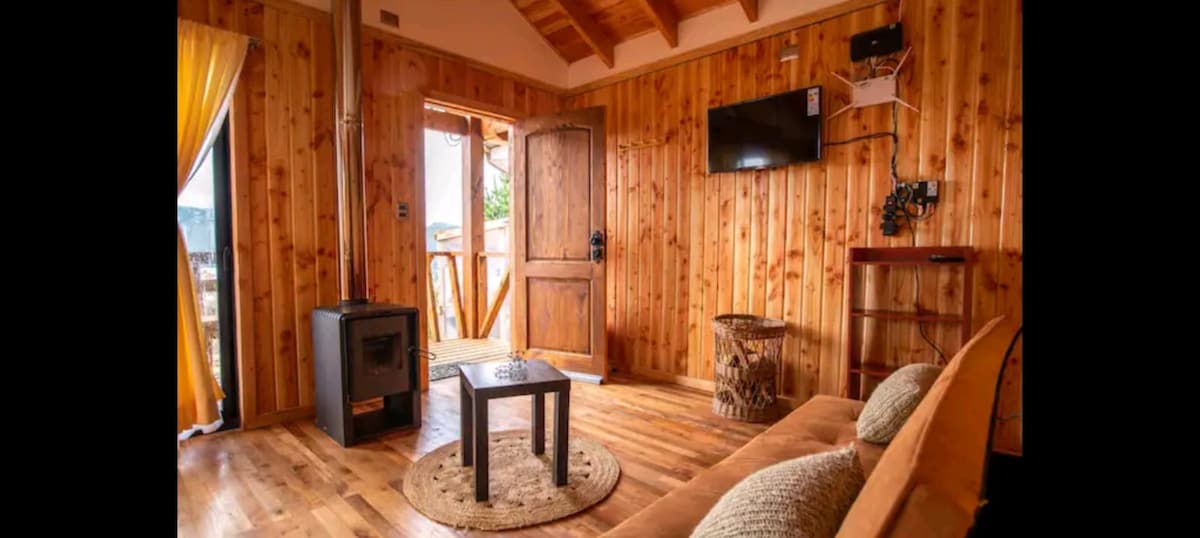 A warm living area features wooden walls and a rustic ceiling. A comfortable sofa sits opposite a small table placed on a round natural fiber rug. A television is mounted on the wall, and a door opens to the outside, inviting natural light.