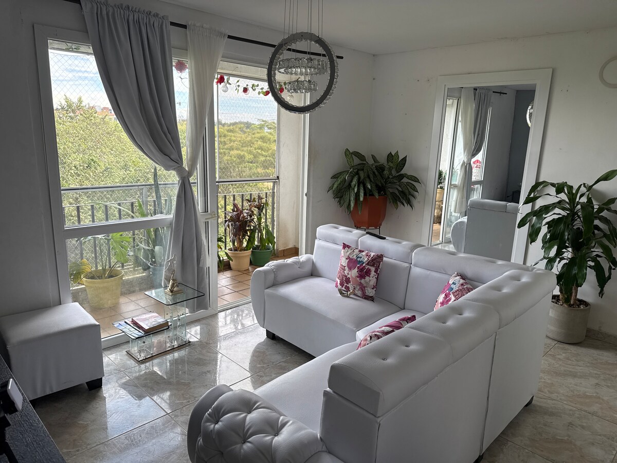 A bright living area features a white sectional sofa adorned with floral cushions. A glass coffee table sits in front, reflecting light from the nearby floor-to-ceiling windows. Lush potted plants add greenery, while a decorative chandelier is mounted above.