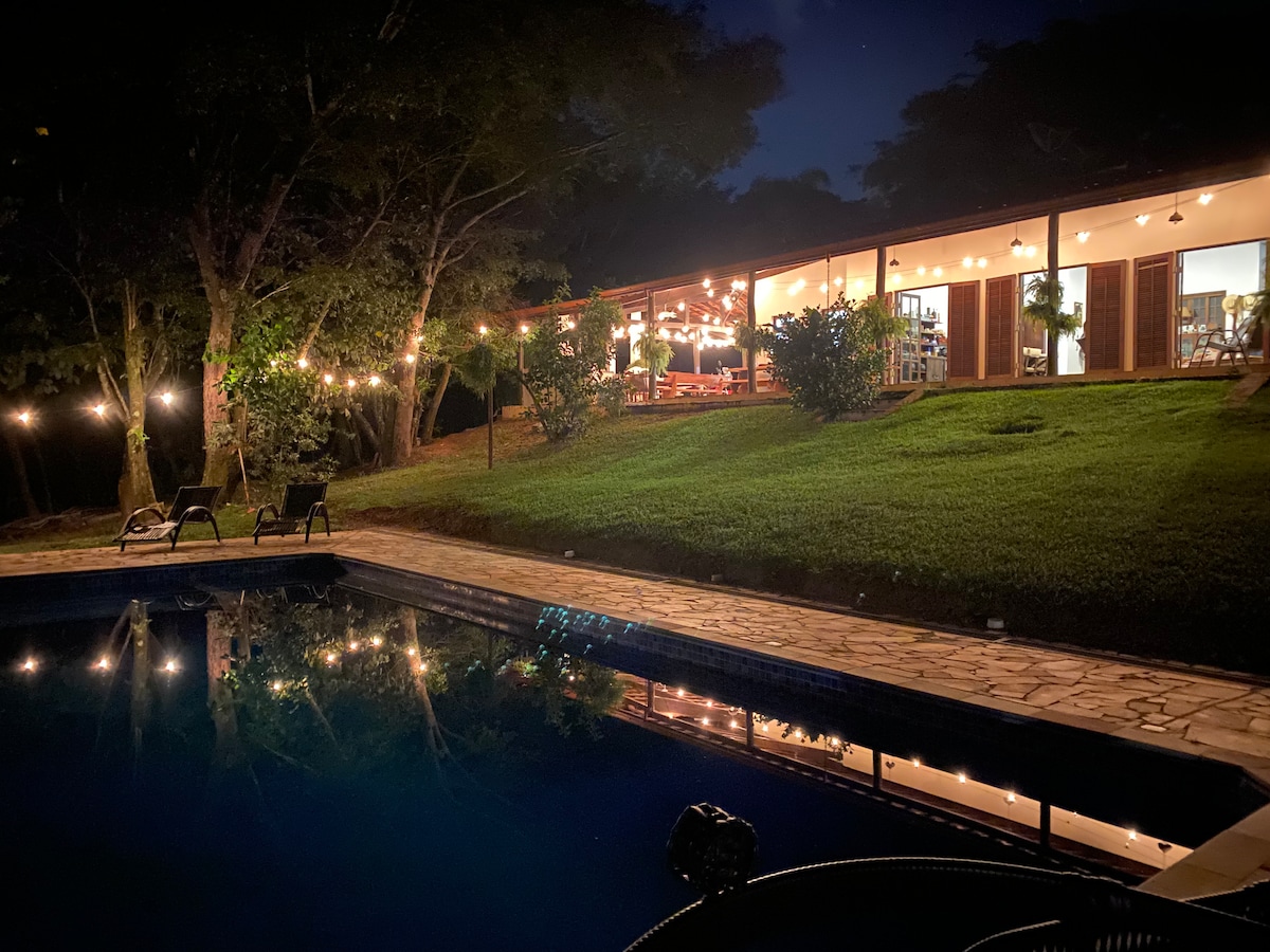 The serene outdoor area features a well-maintained pool reflecting the string lights above. A spacious porch is visible with multiple seating arrangements, surrounded by lush greenery and trees. The warm glow from the lights creates a welcoming ambiance for relaxing evening gatherings.