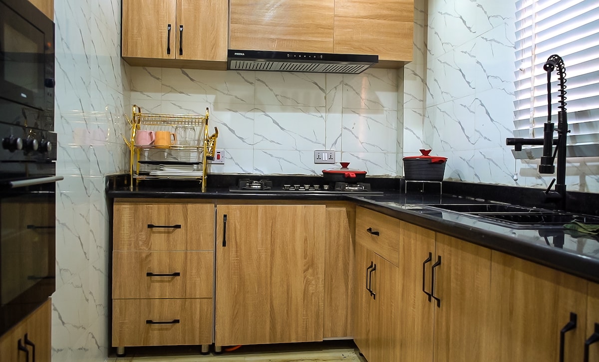 A modern kitchen features wood cabinetry with black hardware, complemented by sleek black countertops. Essential appliances, including a stove, oven, and sink, are visible. A dish drying rack is placed on the counter, with organized dishware nearby. Natural light streams through a partially opened window.