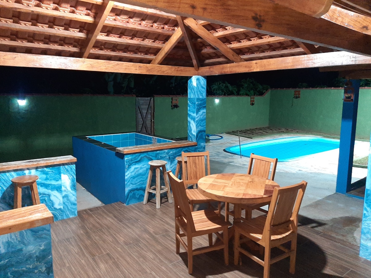 An outdoor area features a wooden table and several chairs, surrounded by vibrant blue columns. A small jacuzzi is visible nearby, while a swimming pool takes center stage in the background, illuminated softly by ambient lighting amidst lush greenery.