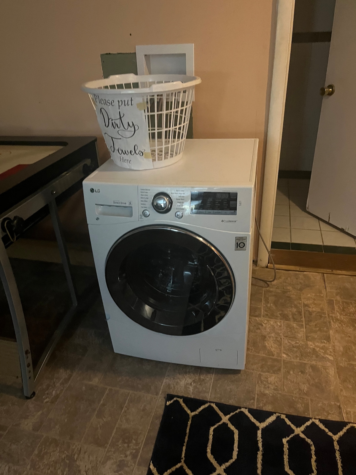 A laundry area features a front-loading washer with a user-friendly control panel. A woven laundry basket sits atop the machine, labeled for easy identification of laundry placement. The flooring consists of textured tiles, and a door is visible in the background.