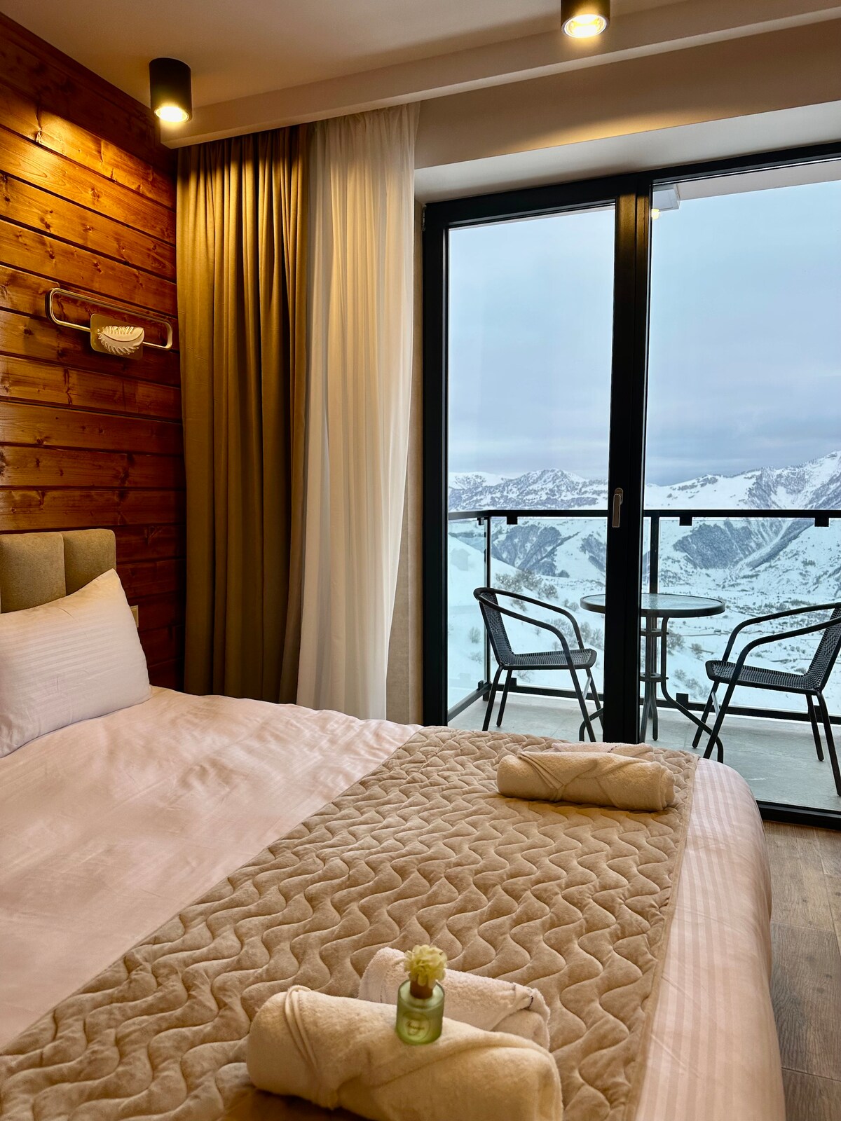 A modern sleeping area is displayed, featuring a spacious bed covered with a light quilt. Pillows are arranged neatly on top. A door leads to a balcony with two chairs and a small table, offering views of snowy mountains in the background.