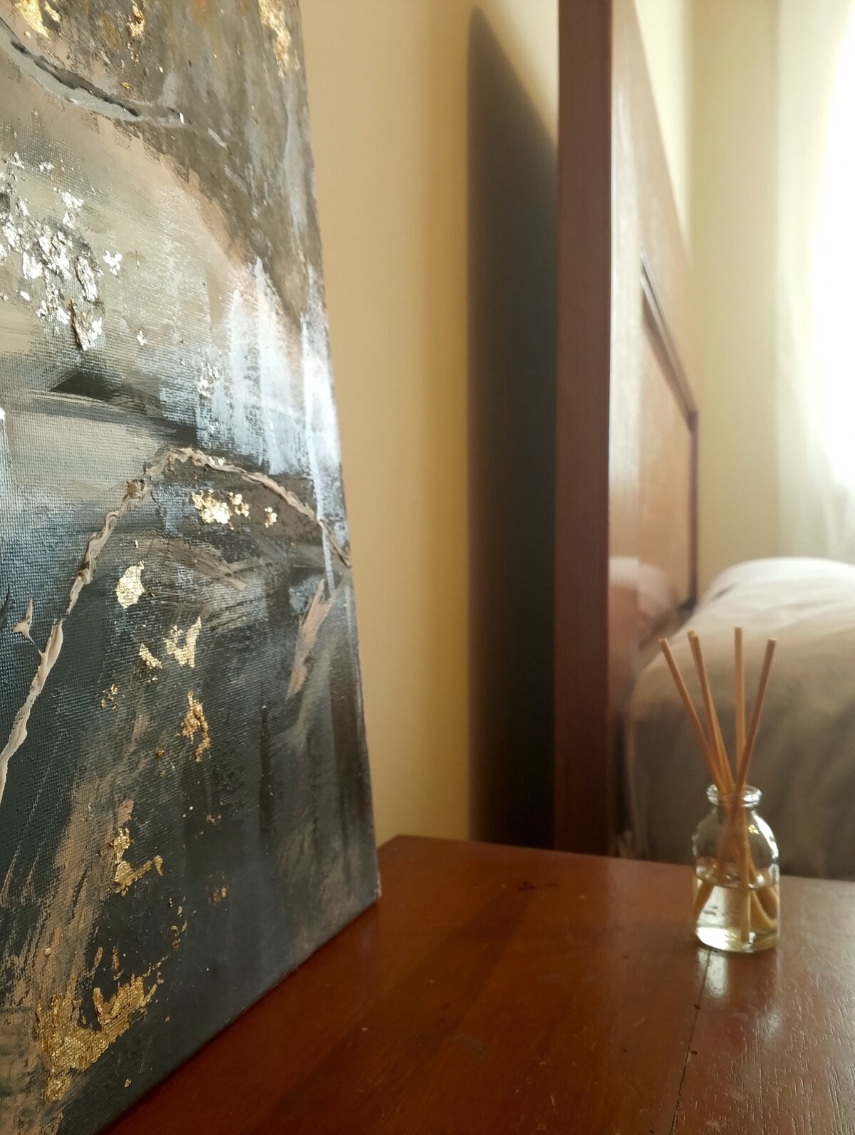 A close-up view of a textured painting featuring abstract designs in muted tones, accented with gold leaf. A small glass jar containing decorative sticks is positioned on a wooden table, with a comfortable bed partially visible in the background, illuminated by natural light from a nearby window.