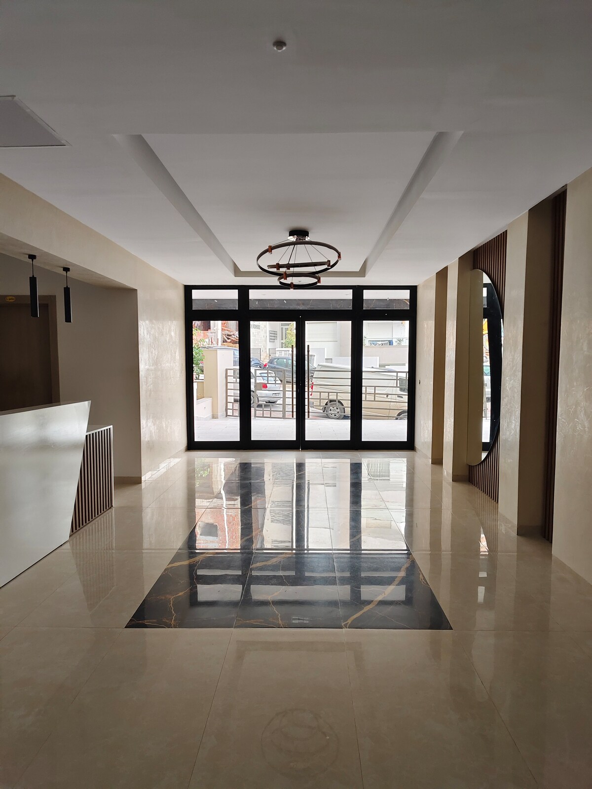 A well-lit entrance hall features elegant flooring with contrasting tiles, including a polished black section. Large glass doors provide exterior views, while modern light fixtures hang from the ceiling. A sleek reception area is visible to the left, adding to the contemporary ambiance.