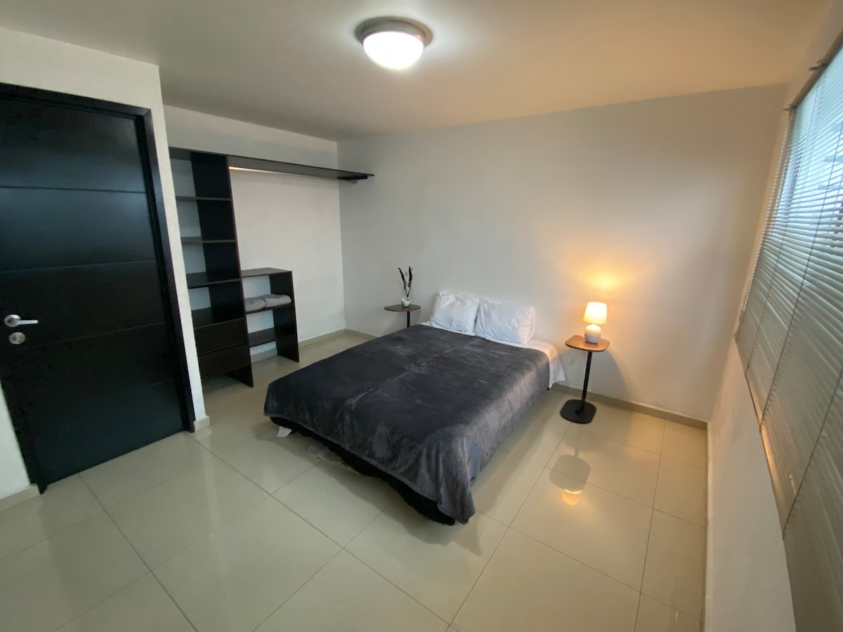 A well-lit bedroom includes a spacious bed with a dark blanket. A bedside lamp provides soft illumination, while a small table sits next to the bed. Shelving is mounted on the wall opposite a window, which allows natural light to enter the room.