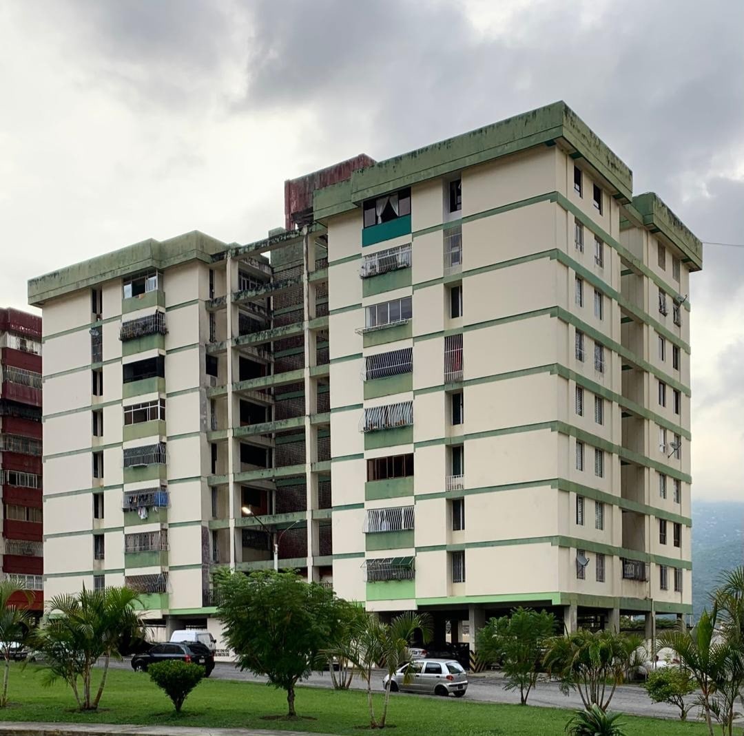 A multi-story building is presented, showcasing a modern architectural design with a light-colored exterior. The structure features several balconies and large windows. Surrounding greenery and a spacious outdoor area contribute to the residential environment. Parking is visible at the base of the building.