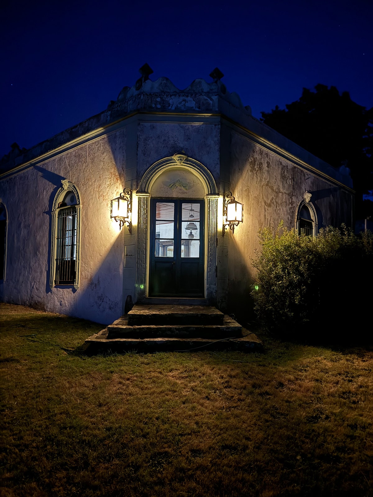 The exterior of the historic house is illuminated by lanterns at night, showcasing intricately designed arches and windows. A stone staircase leads up to the double door entrance, framed by a well-maintained lawn and shrubs providing a serene and inviting ambiance.