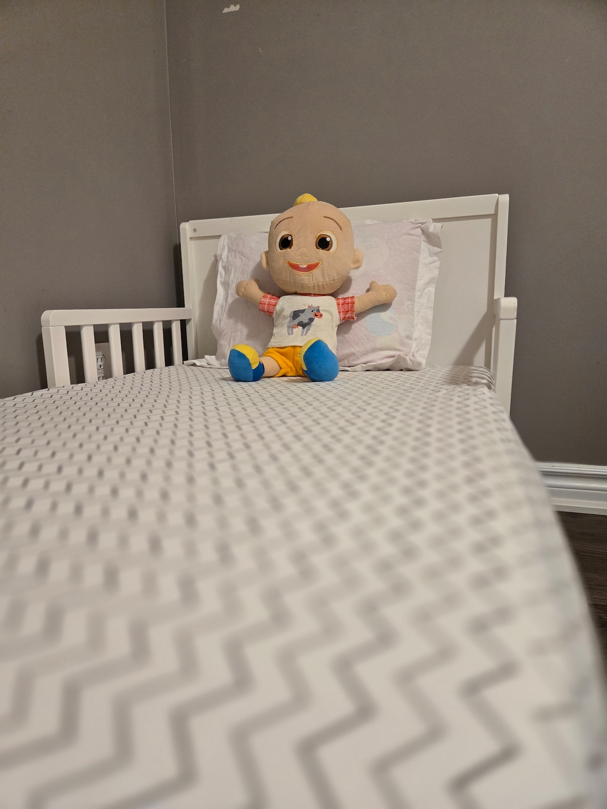 A toddler bed is positioned against a gray wall, adorned with a simple zigzag-patterned bedspread. A cheerful plush toy sits on the pillow, introducing a playful element to the space.