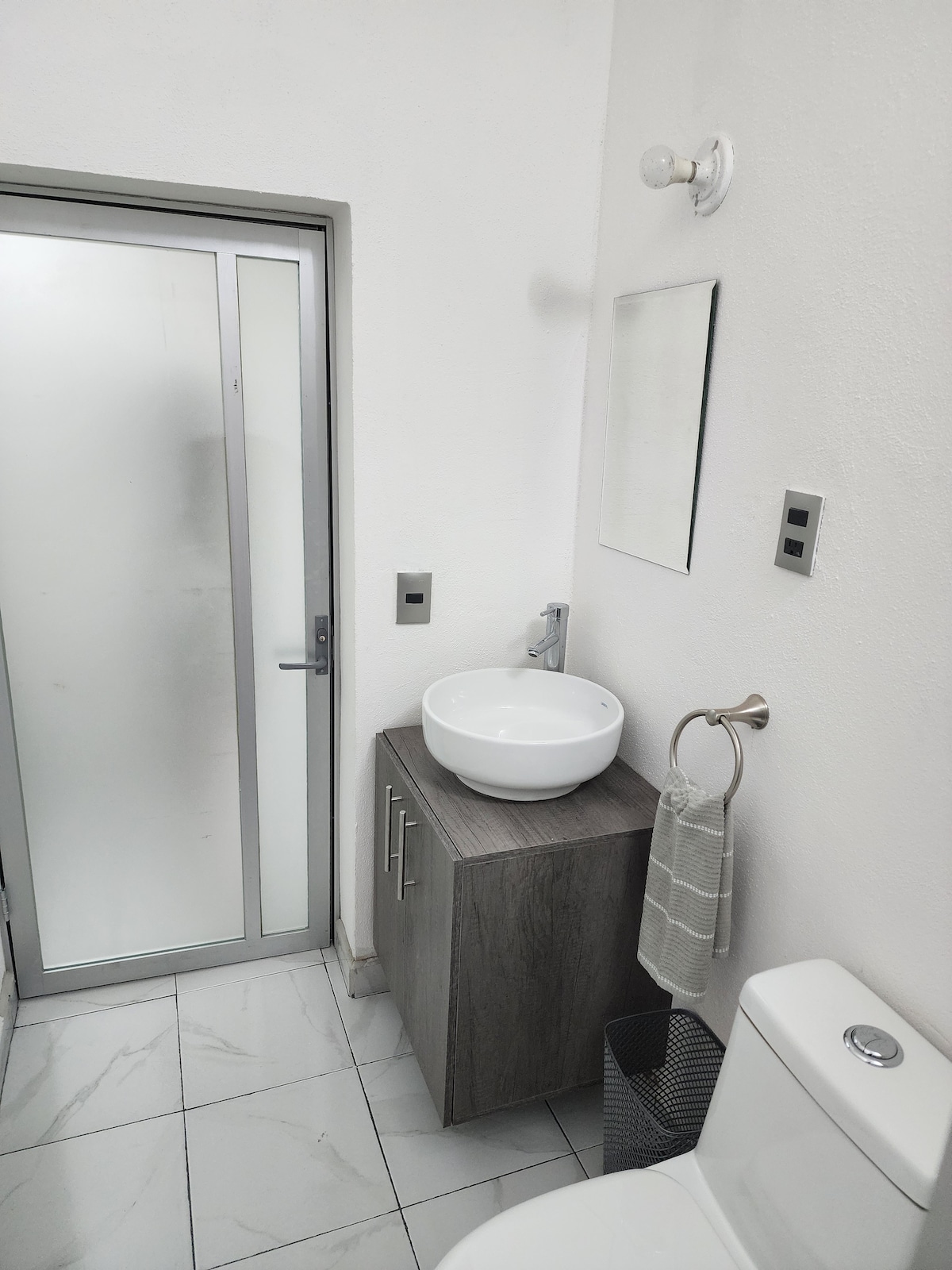 A clean and modern bathroom features a frosted glass shower enclosure, a minimalist sink bowl set on a gray cabinet, and a light fixture above. A towel rack with a gray towel and a wastebasket complete the space, which is highlighted by white walls and tiled flooring.