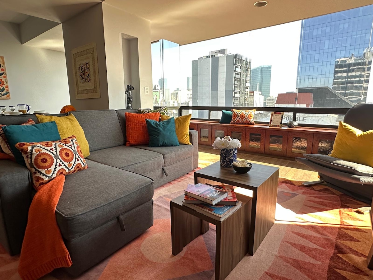 A comfortable living area features a gray sofa adorned with colorful pillows. A small wooden table sits on a patterned rug. Large windows overlook the cityscape, allowing ample natural light to enhance the ambiance.