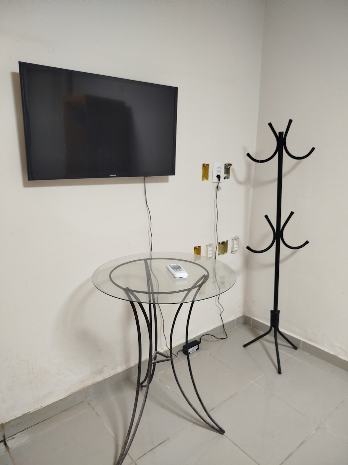 A wall-mounted television is displayed above a glass side table with a remote control placed on it. A black coat rack stands nearby, complementing the light-colored walls and tiled floor.