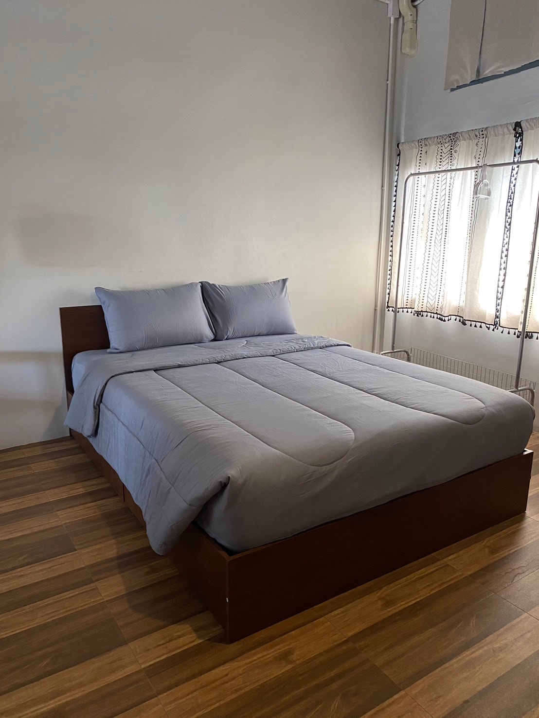 A spacious bed is dressed in soft grey linens, complemented by matching pillows. Natural light enters through a nearby window, which is partially adorned with light sheer curtains. The polished wooden floor adds a warm touch to the serene sleeping area.