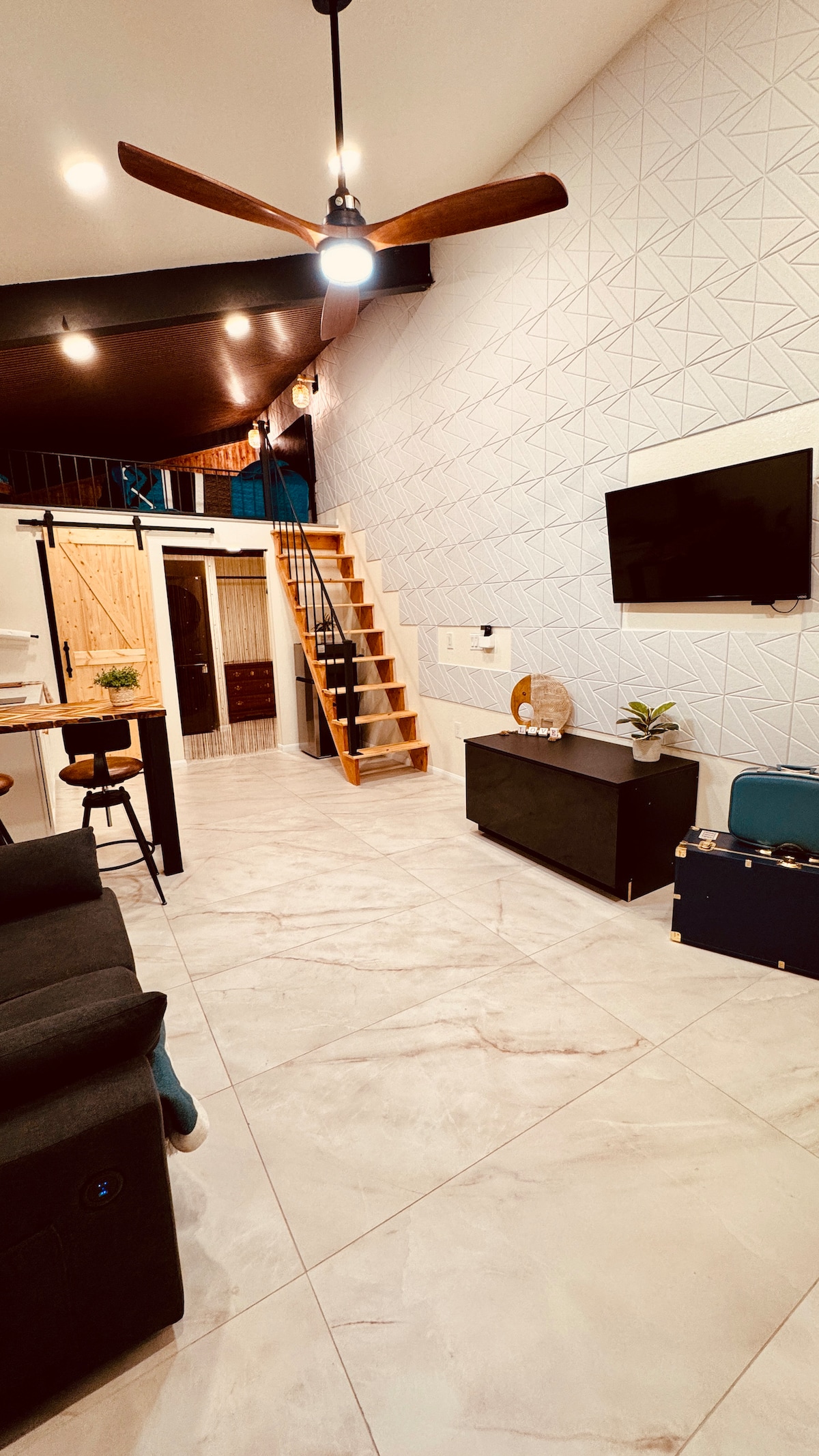 An open-concept living area is presented, featuring a sleek black sofa and a mounted flat-screen television. A wooden staircase leads to an upper level. Soft lighting illuminates the geometric-patterned wall, while a small plant and decorative items are arranged on the dark media console.