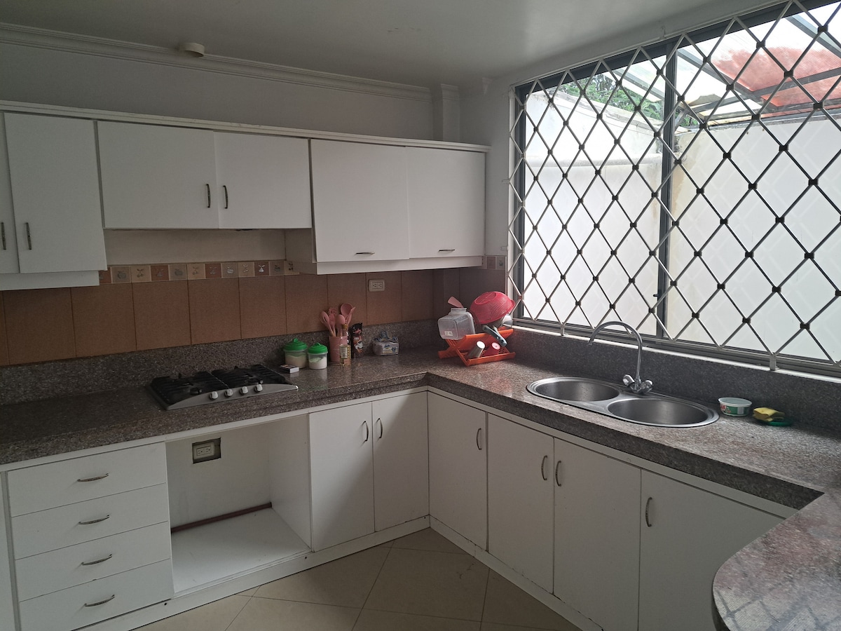 A well-equipped kitchen features a modern layout with white cabinetry and a granite countertop. Two stainless steel sinks sit under a large window, offering natural light. A gas stove is positioned on the countertop, complemented by various kitchen essentials organized nearby.