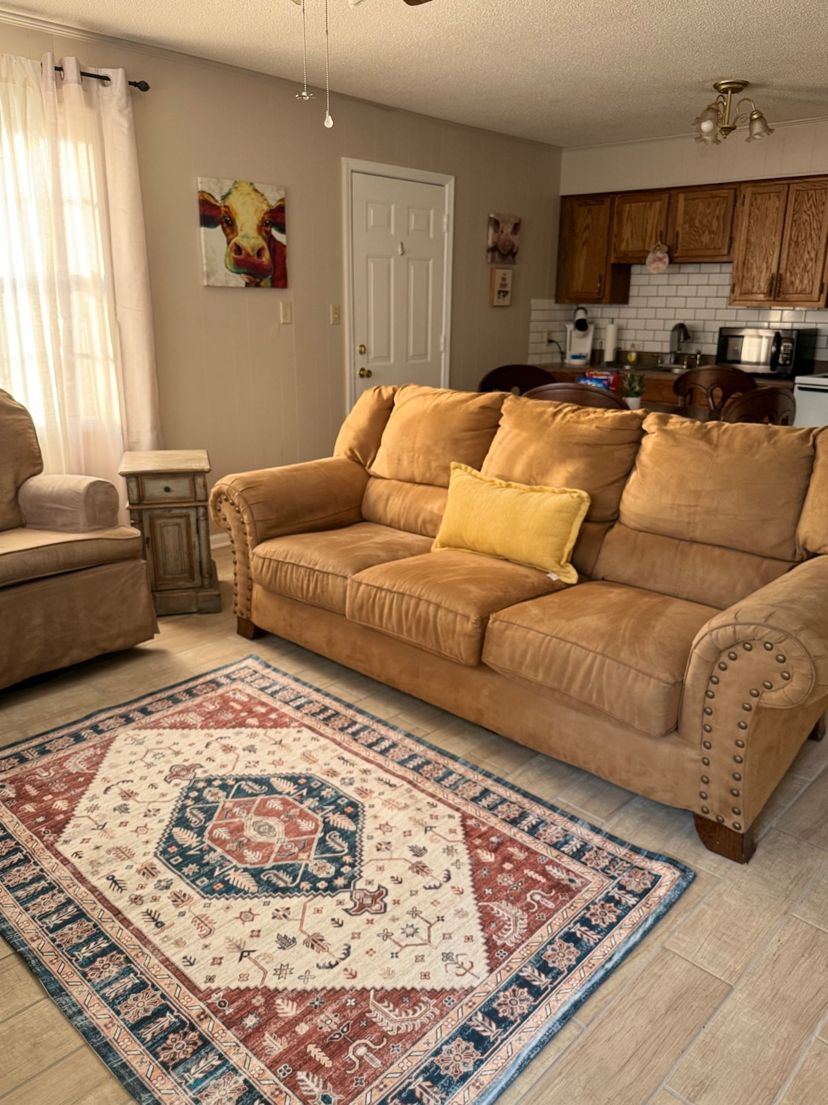 A comfortable living area features a plush, light brown sofa with decorative nailhead trim and a yellow accent pillow. A traditional area rug with intricate patterns lies beneath, complementing the space, while warm sunlight filters through sheer curtains, illuminating the cozy atmosphere.