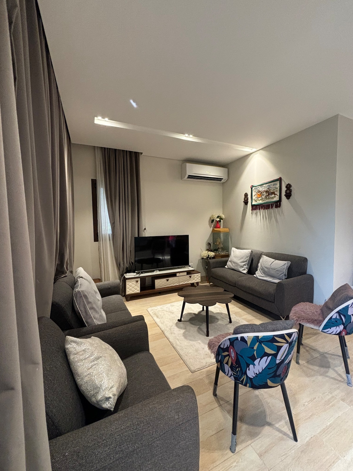 A cozy living area features two dark gray sofas and a patterned chair surrounding a circular coffee table. A television is mounted on the wall, with decorative items placed nearby. Soft lighting is provided by recessed ceiling lights, and curtains gently frame the windows.