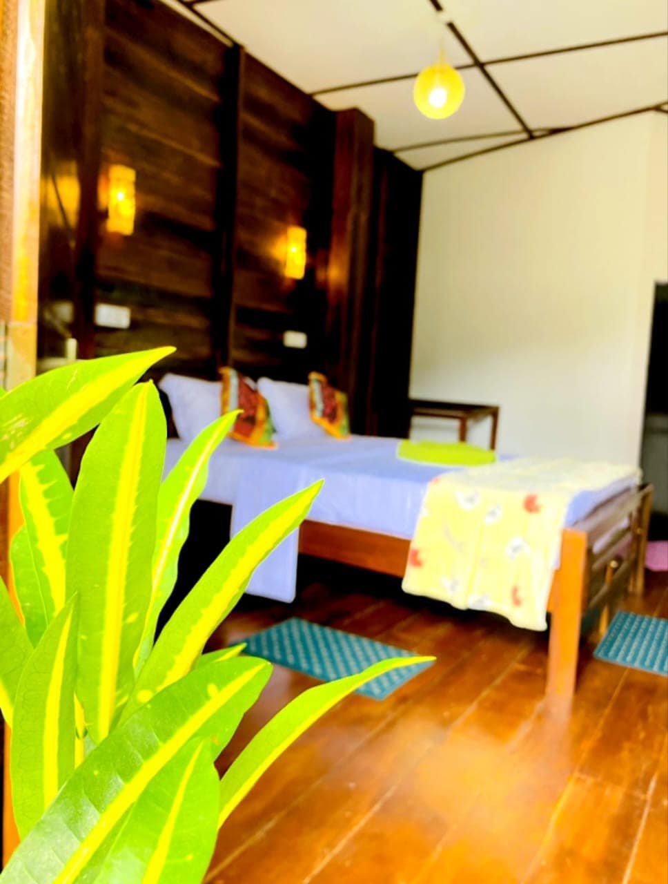 A serene bedroom space is visible, featuring a wooden bed topped with colorful pillows and a light blanket. Natural light filters through the room, complemented by wooden paneling and soft wall lights. In the foreground, green leaves provide a touch of nature.