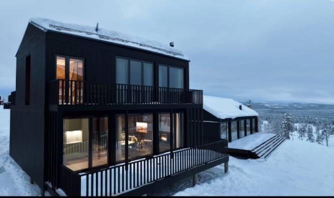 The exterior of the villa is shown against a serene winter landscape, featuring dark wood siding and large glass windows. A spacious deck with horizontal railings encompasses the front, while snow-covered surroundings highlight the scenic mountain views.