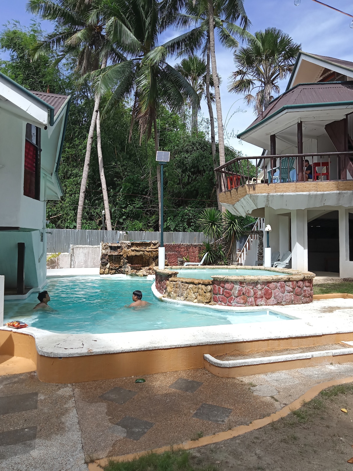 A spacious outdoor area is featured, showcasing a swimming pool with clear blue water and a stone spa. Lush palm trees surround the space, providing a sense of privacy. The two-story building offers multiple balconies overlooking the pool area.