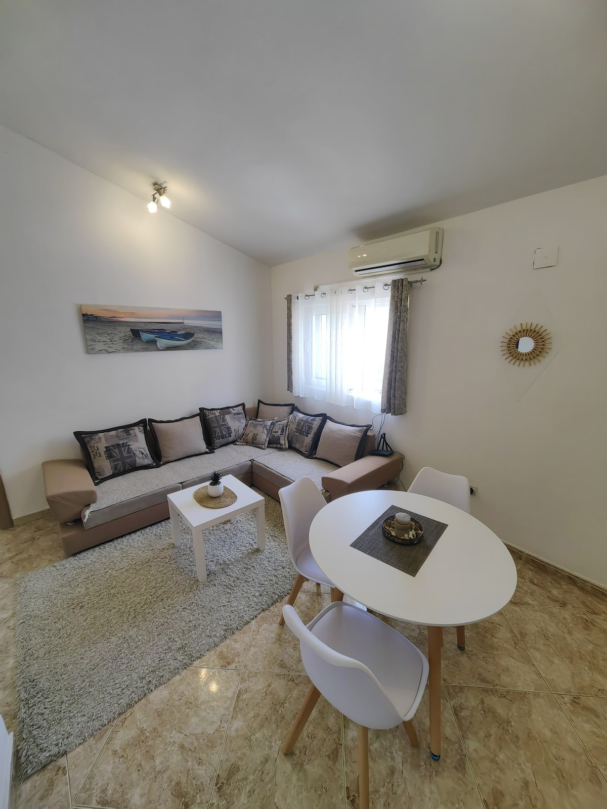 A well-arranged living area features a comfortable sofa with decorative cushions and a white coffee table on a soft area rug. A small dining table with four chairs offers seating. Natural light filters through the sheer curtains adorning the window, enhancing the warm ambiance.