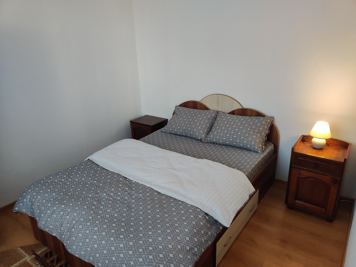 A cozy bedroom features a spacious bed with a patterned gray duvet and two pillows. A soft light emanates from a bedside lamp, illuminating a wooden nightstand on one side. Light wood flooring extends throughout the area, creating a simple and welcoming environment.