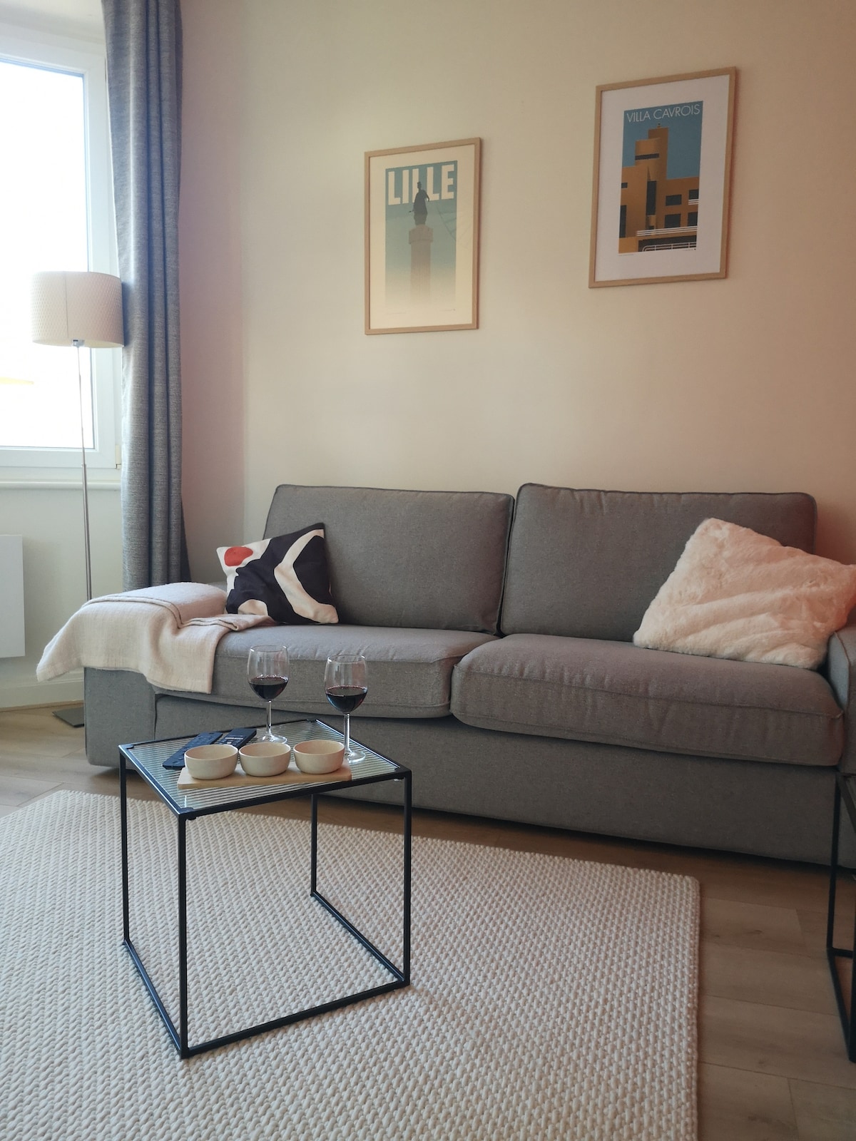 A comfortable gray sofa is paired with a cozy throw and decorative pillows. A small coffee table, topped with two glasses and snacks, is positioned in front. Art prints adorn the wall above, and natural light enters through the nearby window.