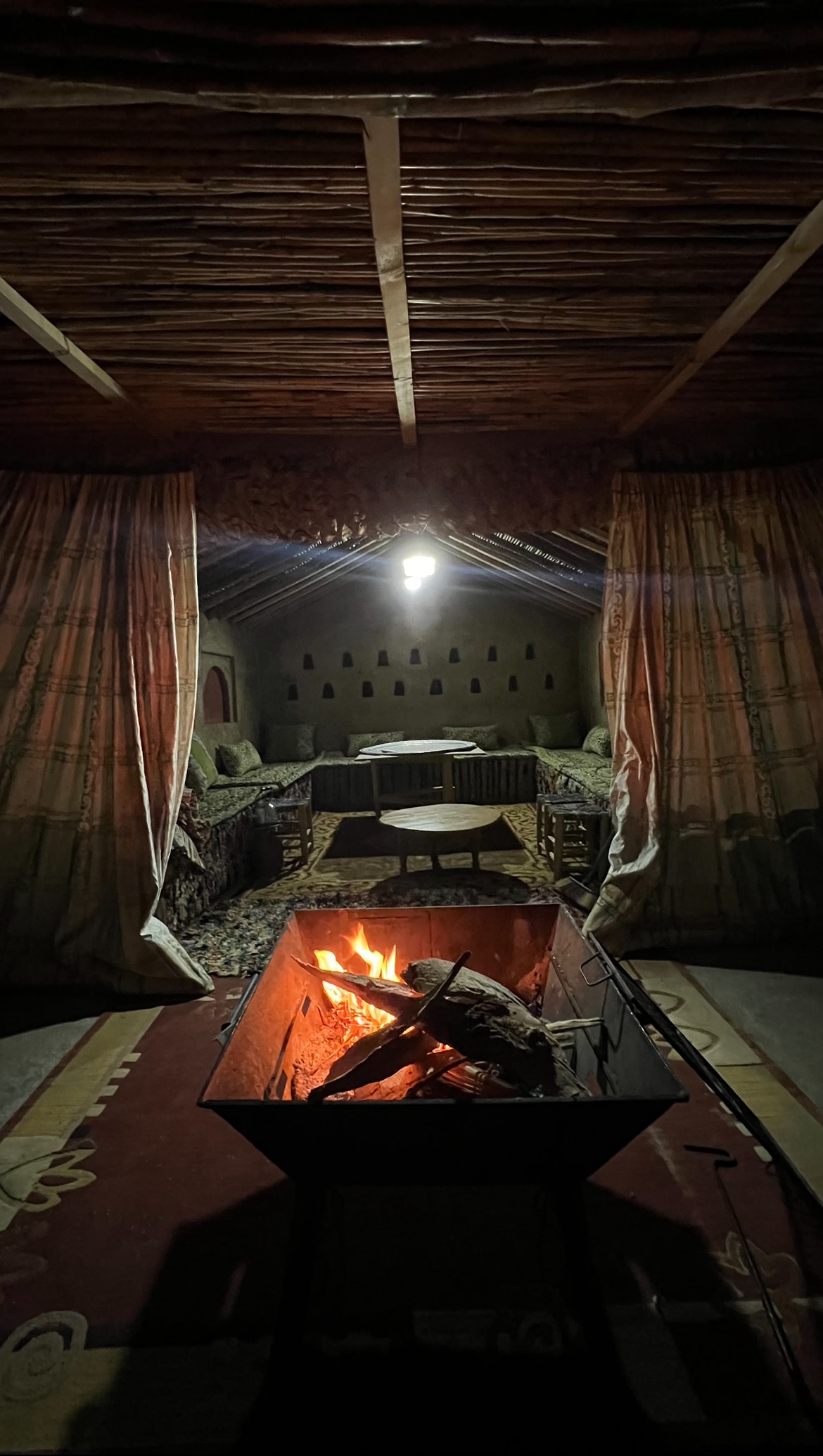 A cozy sitting area is featured, with plush seating surrounding a central fire pit. Warm light from a hanging bulb illuminates the space, while patterned curtains frame the entrance. The backdrop reveals additional seating areas, creating an inviting atmosphere for gatherings.