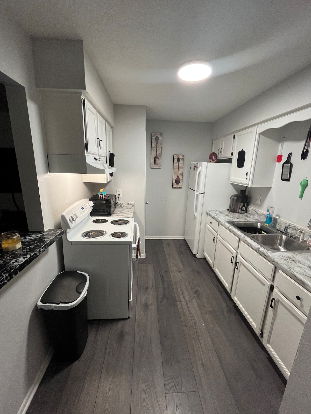 A functional kitchen is equipped with essential appliances including a stove, refrigerator, and sink. White cabinetry and a dark marble countertop create a clean look. The floor features dark wood, and the space is illuminated by a ceiling light, enhancing visibility.