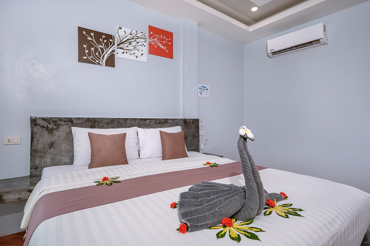 A spacious bed is adorned with decorative pillows and a subtle floral arrangement. Elegant towel art shaped like a swan is arranged on the covers, while soft wall art adds a touch of creativity. The light blue walls create a calming backdrop, complemented by an air conditioning unit.