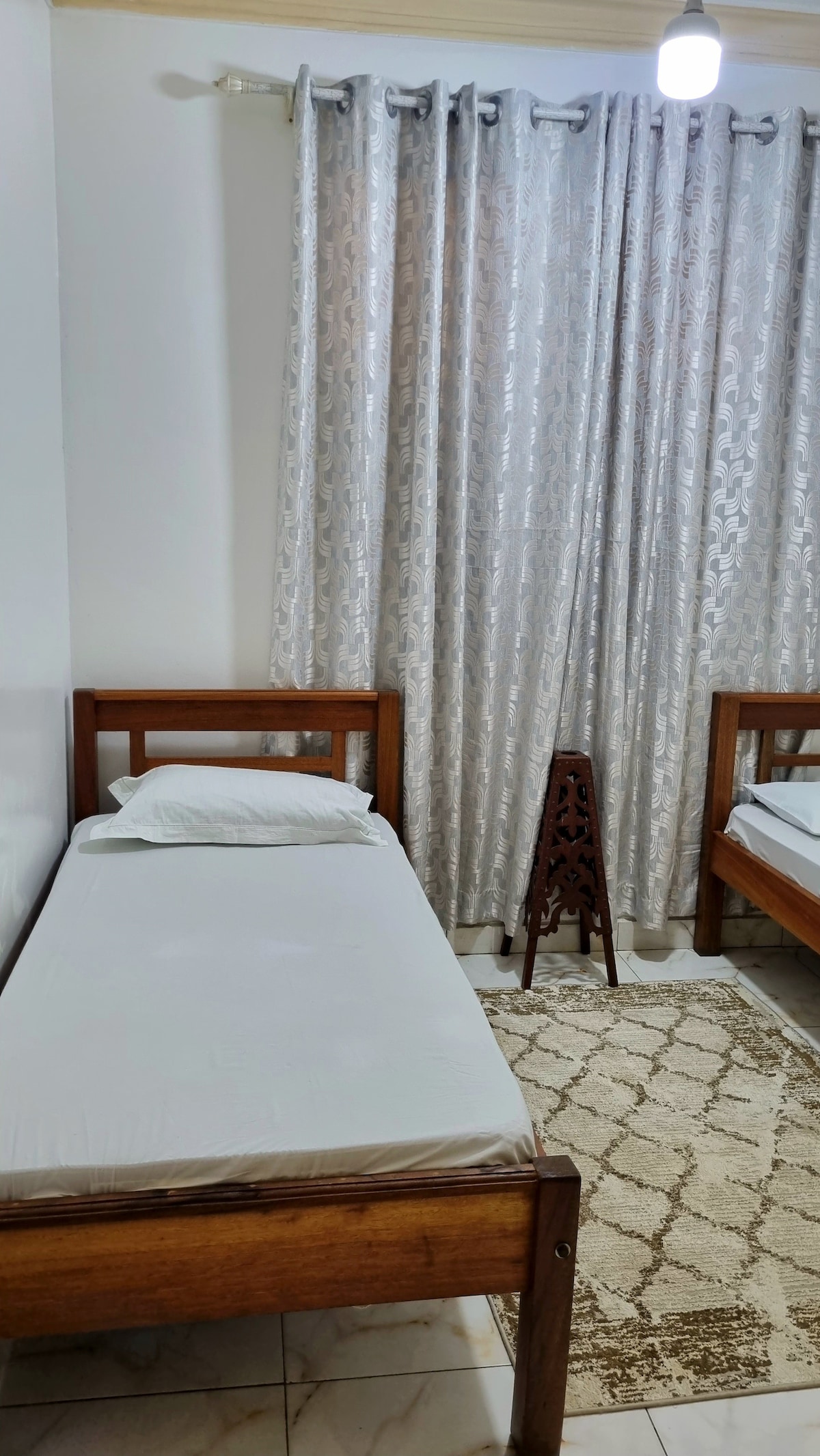 A sleeping area features two wooden single beds positioned beside each other. Soft bedding is neatly arranged on each bed. A light-colored rug lies on the tiled floor, complemented by a decorative wooden stand. Sheer curtains gently filter sunlight from a nearby window.