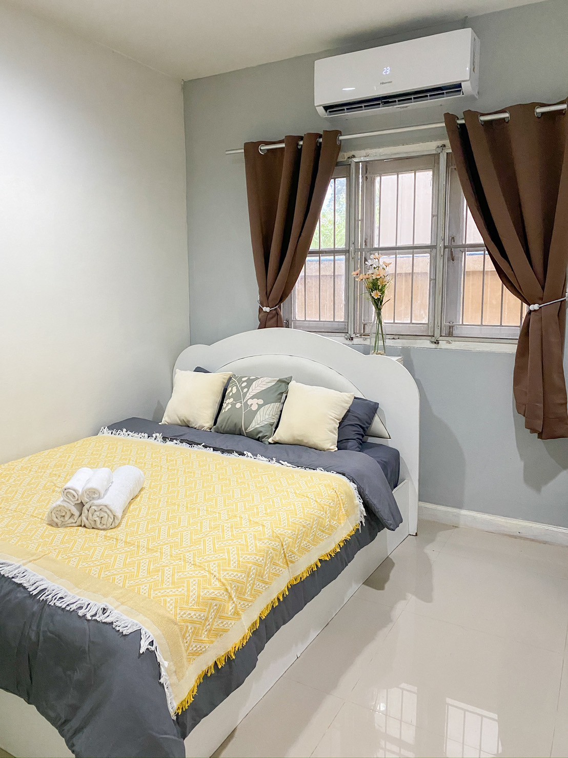 A compact bedroom features a spacious bed adorned with a yellow geometric blanket and decorative pillows. Natural light enters through a window dressed with brown curtains. A vase with flowers sits on the bedside, while air conditioning is mounted above for comfort.