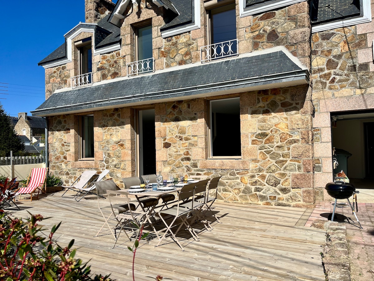 The outdoor terrace features wooden decking furnished with a dining table and chairs, suitable for gathering. A grill is positioned nearby, surrounded by natural stone walls of the house. Several lounge chairs are placed at one side, offering a comfortable seating area under the clear blue sky.